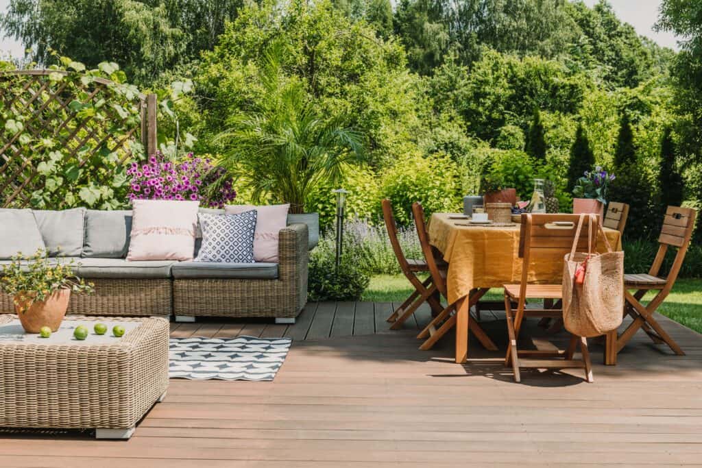 Balcon en bois avec mobilier d'extérieur, canapé en rotin et table à manger dans un jardin verdoyant.
